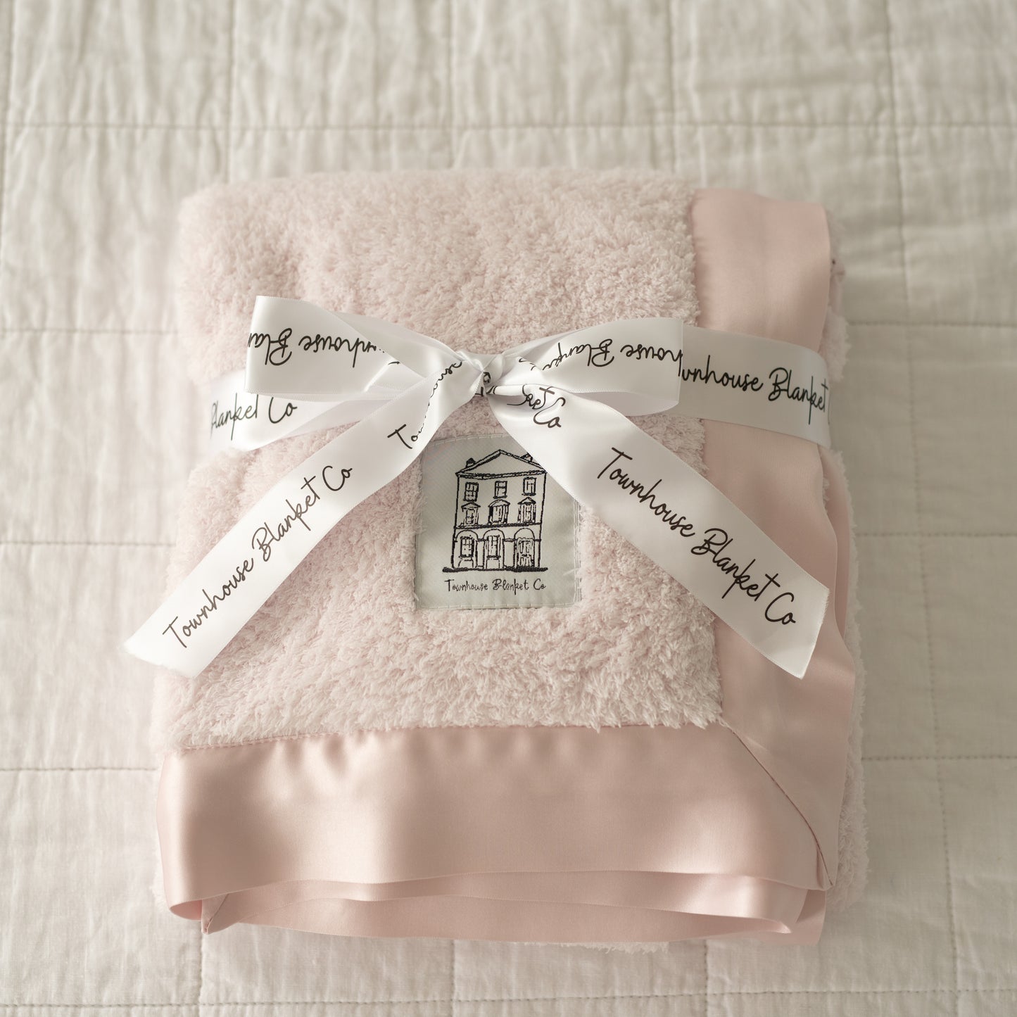 Delicately folded luxury pink Townhouse baby blanket, wrapped in an elegant white ribbon with black Townhouse branding, tied in a classic bow. Featuring an ultra-soft pink satin edge, this exquisite blanket is the perfect keepsake gift for a baby girl.