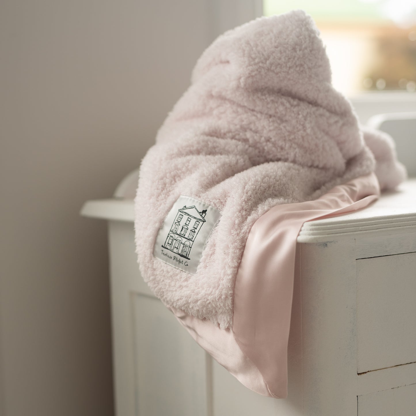 Pink soft chenille Townhouse Blanket Company luxury blanket with pink satin edges and logo patch, draped on a white side table in a boho chic bedroom.