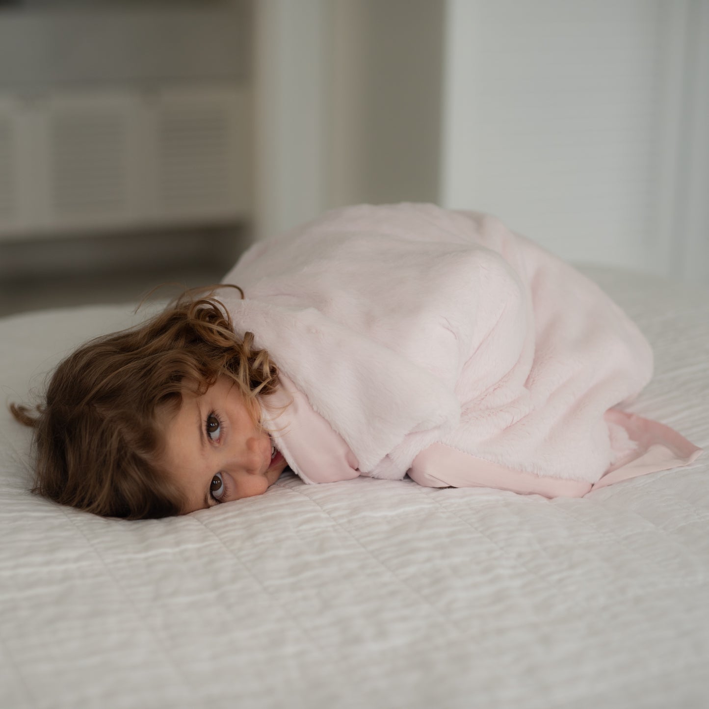 Cute brown-eyed girl lying on bed wrapped in a pink luxury Townhouse Blankets baby blanket with satin edge, hair flowing over back.