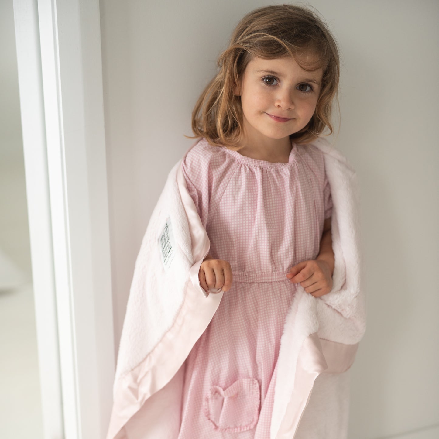 Smiling girl with brown eyes in pink gingham dress holding a super soft luxury pink Townhouse Blanket Company forever blanket with satin edges against a white wall.