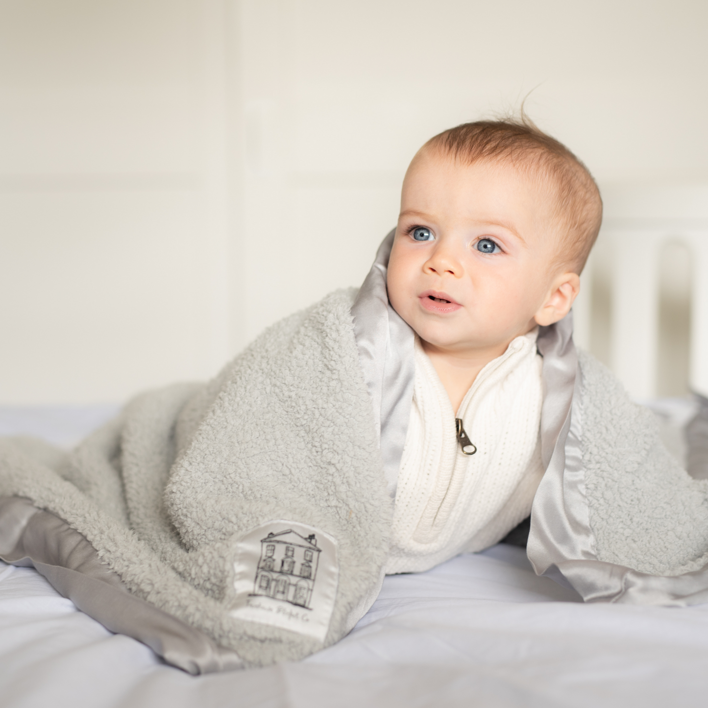 Adorable baby with bright blue eyes crawling on a bed, wrapped in a luxuriously soft grey chenille Townhouse blanket with silky satin edges. The blanket drapes gently over his back as he looks to the left, smiling happily—capturing the joy of ultimate comfort and warmth.