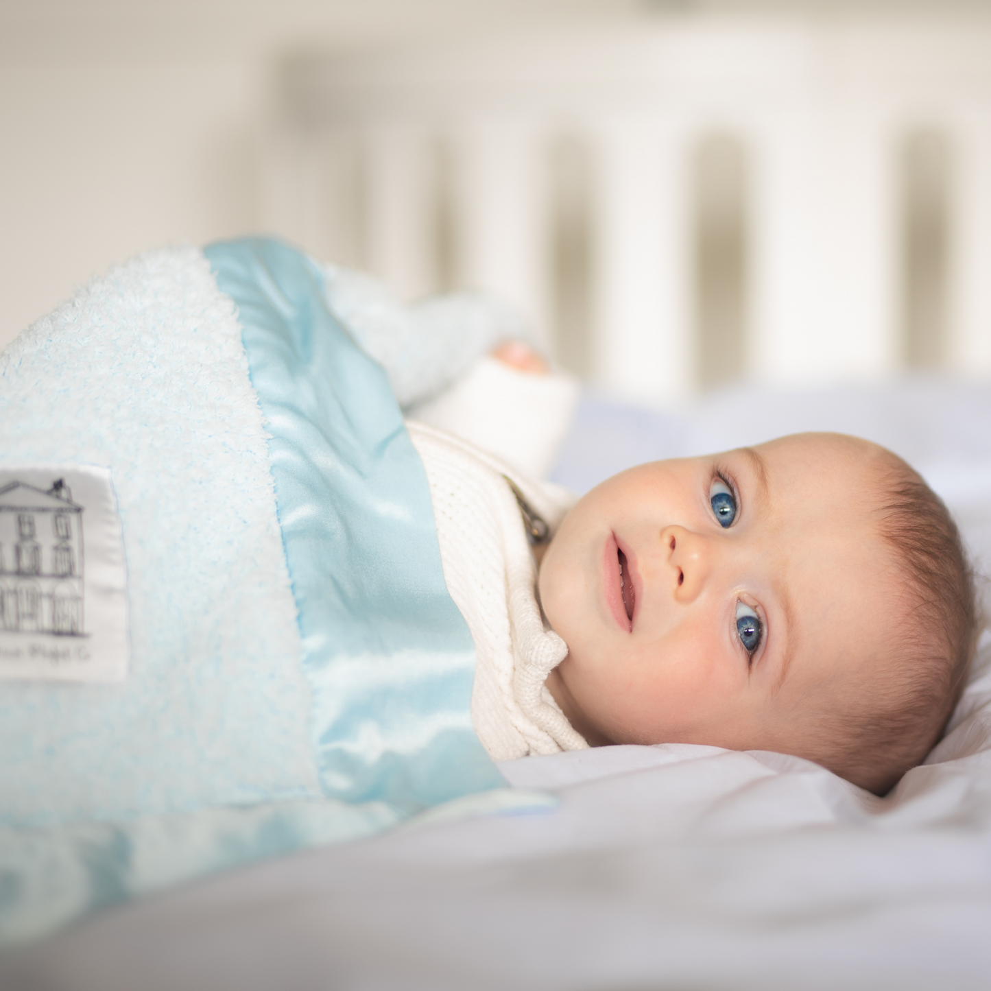 Wide awake blue-eyed baby boy smiling on cot under a blue super soft chenille Townhouse Blankets luxury forever blanket with satin edge and logo patch.