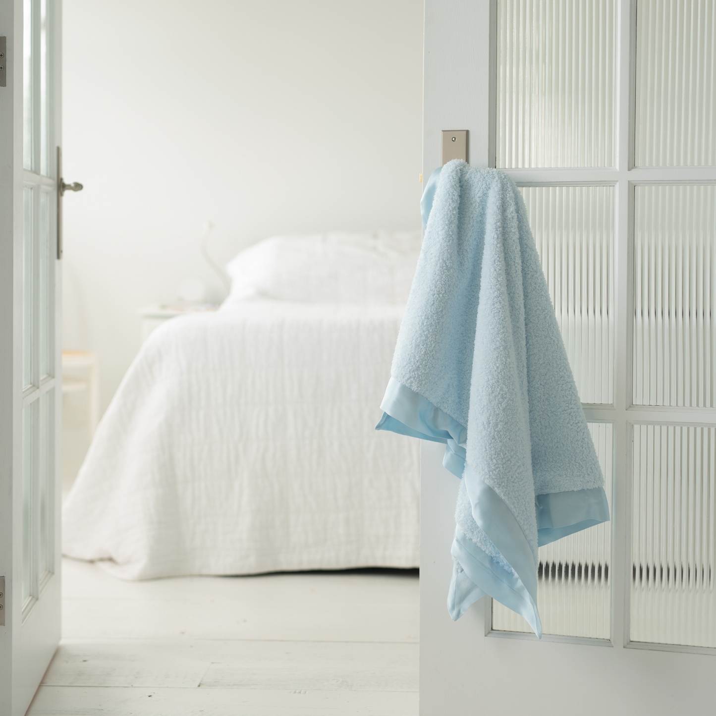 Blue chenille baby blanket with soft satin edges draped over a white vintage-style door handle. The open door reveals a white-covered double bed and white-painted stained wood floorboards, creating a boho-chic, timeless nursery aesthetic.