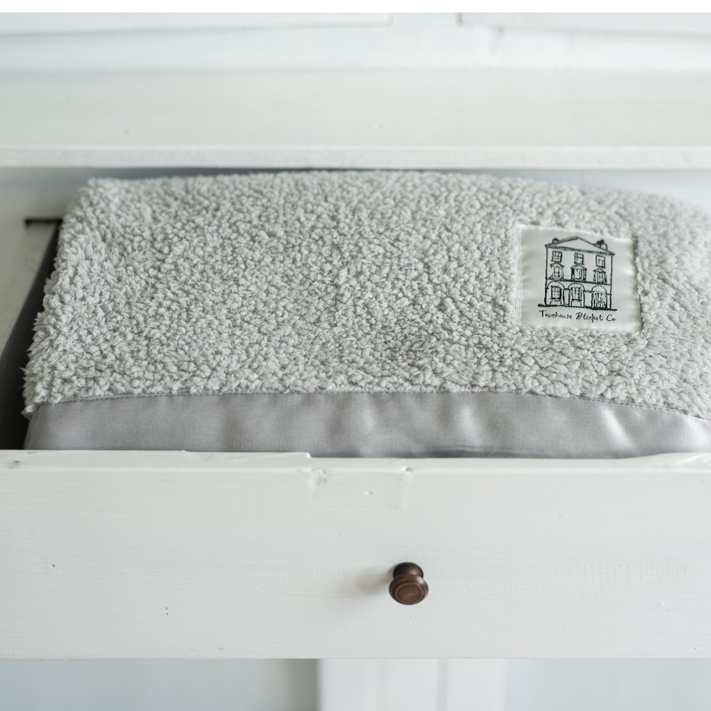 Luxuriously soft grey chenille Townhouse blanket with signature logo patch, neatly folded inside a white chest of drawers. A timeless forever blanket, styled in a boho-chic, shabby-chic setting—perfect for adding warmth and elegance to any home.