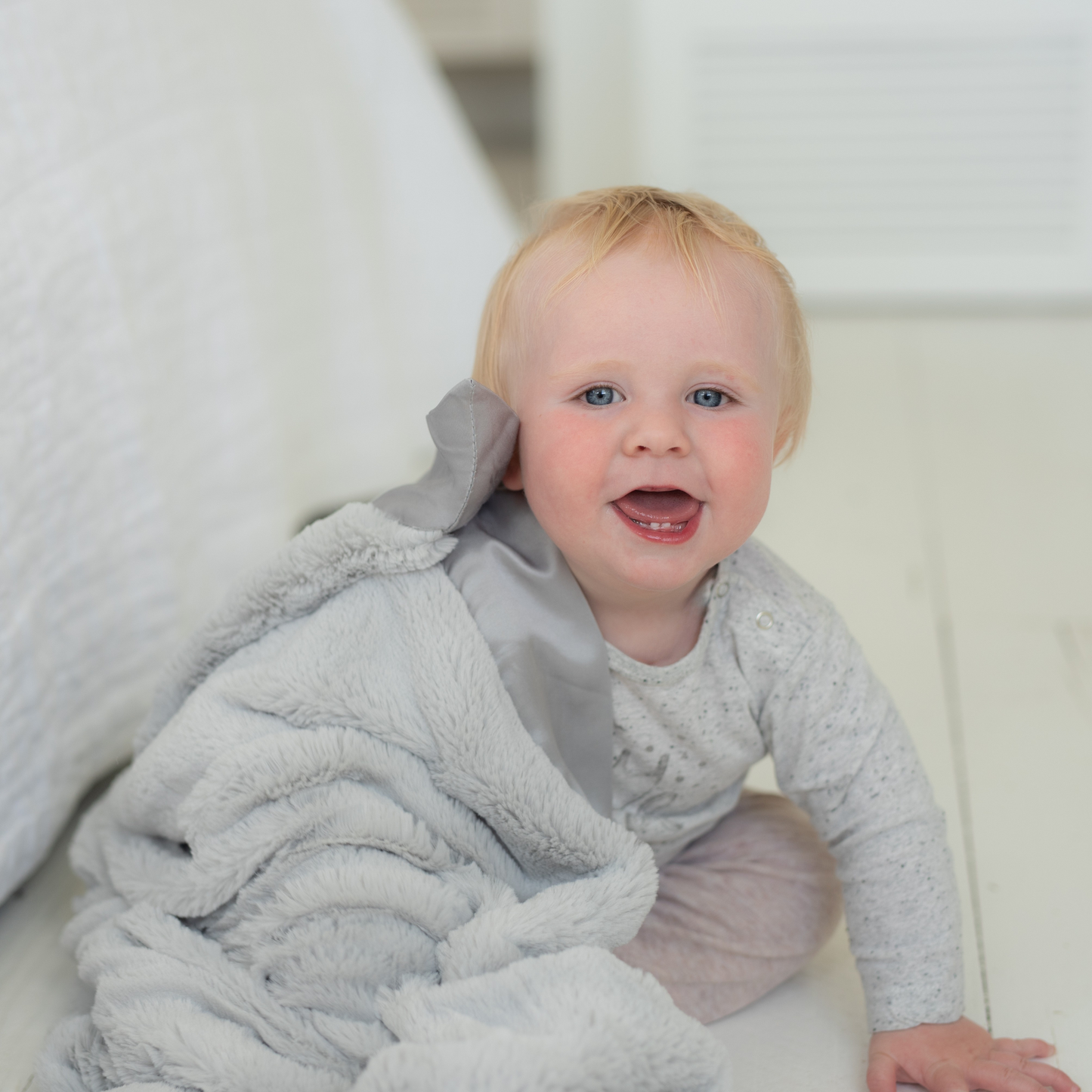 Smiling 1-year-old blue-eyed baby sitting on white wood floor with a luxury grey super soft Townhouse Blanket Company blanket on their shoulder, satin edge by their ear.