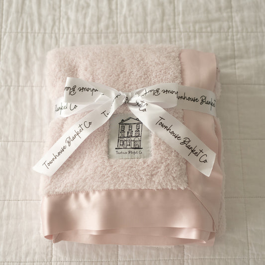 Delicately folded luxury pink Townhouse baby blanket, wrapped in an elegant white ribbon with black Townhouse branding, tied in a classic bow. Featuring an ultra-soft pink satin edge, this exquisite blanket is the perfect keepsake gift for a baby girl.