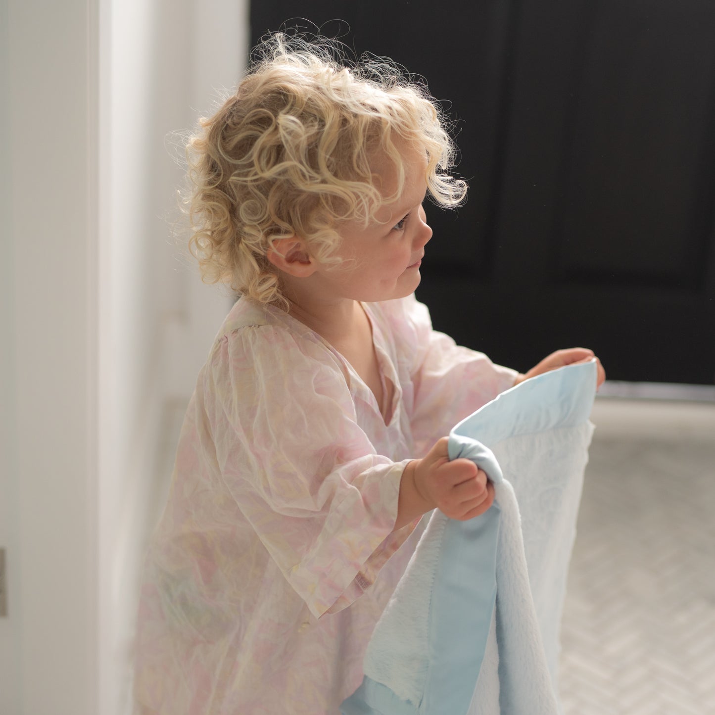 Close-up of an adorable blonde baby with soft curls, snuggled in a luxurious blue blanket with a cool satin edge. The baby’s big blue eyes shine as they hold onto the silky trim, highlighting the blanket’s comforting softness.